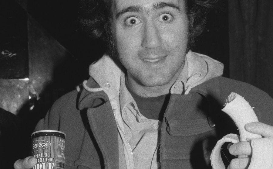 Andy Kaufman is making a goofy expression as he eats a banana and drinks canned apple juice.