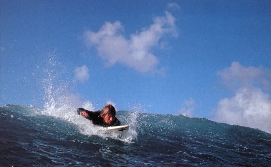 Patrick Swayze, as Bodhi, is surfing the waves. 