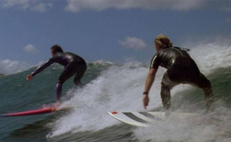 Swayze and Reeves are surfing a wave. 