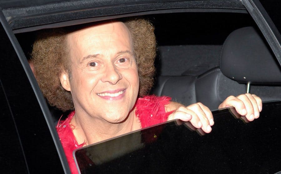 Richard Simmons smiles from a window at the backseat of a car.