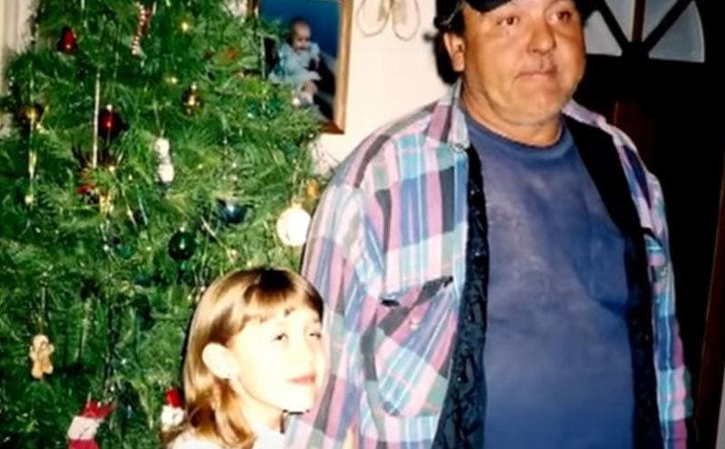Jennifer holds her fathers’ arm as they stand next to a Christmas tree.