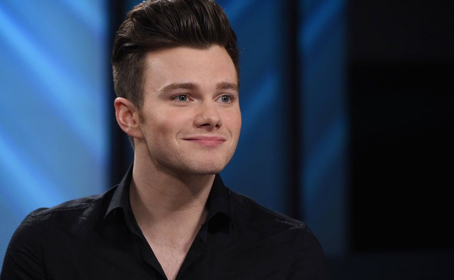 A close up shot of Chris Colfer in a black buttoned up shirt.