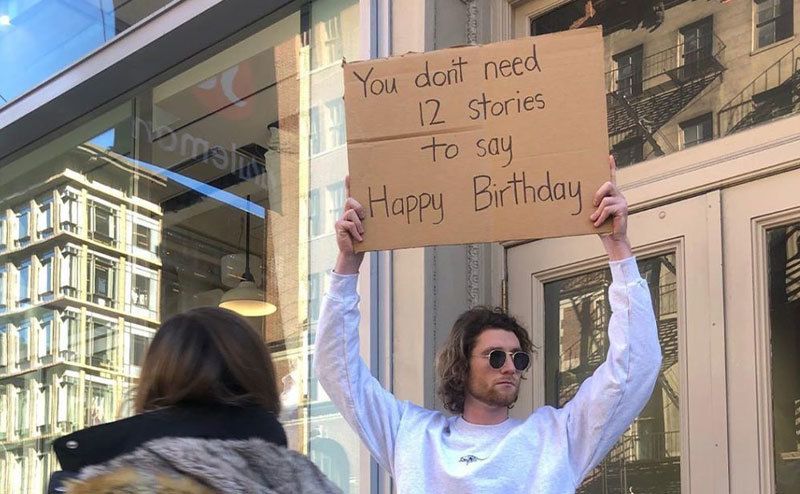 A man is holding a sign that reads ‘’You Don’t Need 12 Stories to Say Happy Birthday.’’
