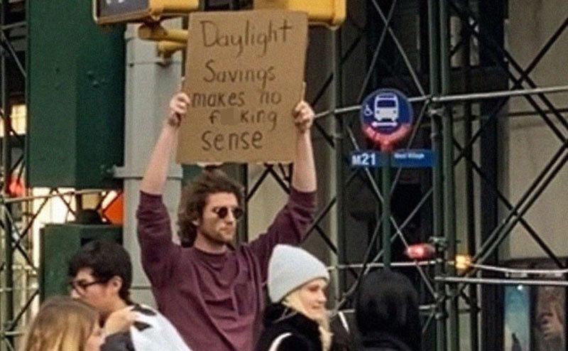 A man is holding a sign that reads ‘’Daylight Savings Makes No Sense.’’