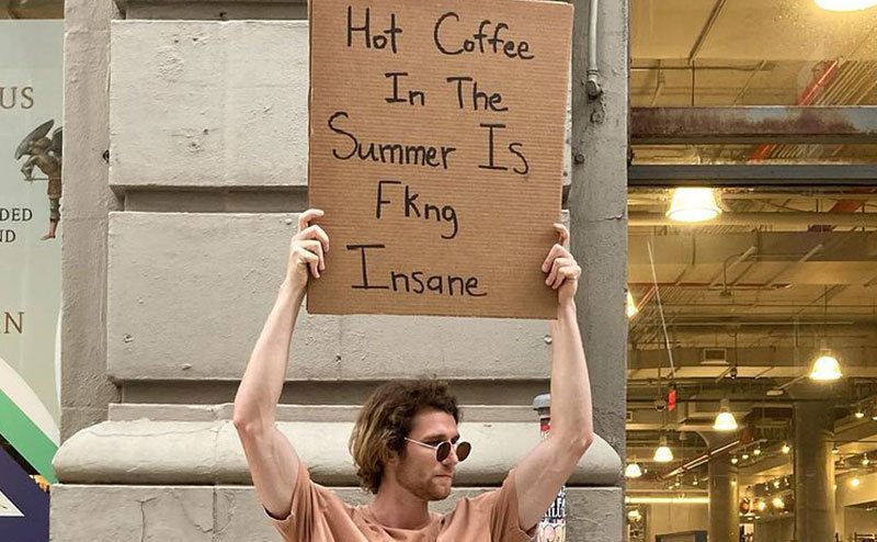 A man is holding a sign that reads ‘’Hot Coffee in this Summer is Insane.’’