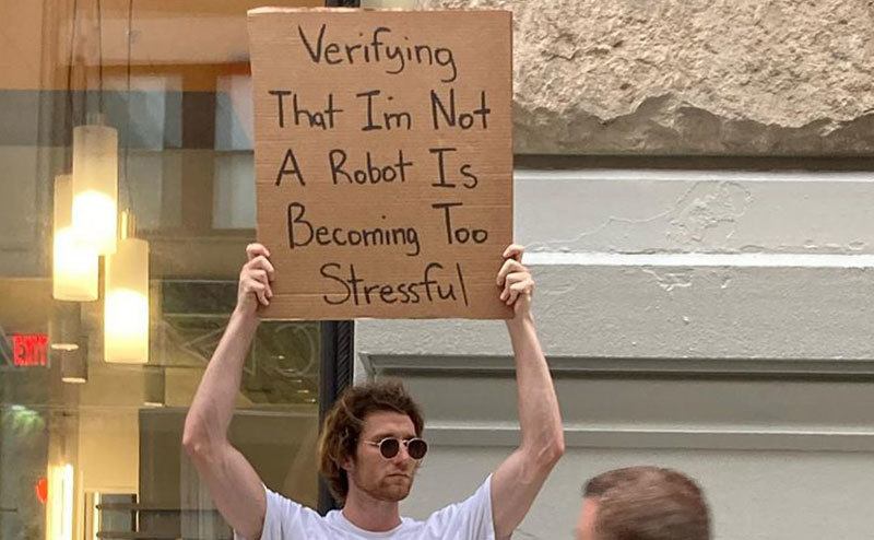 A man is holding a sign that reads ‘’Verifying that I’m not a Robot is Becoming too Stressful.’’