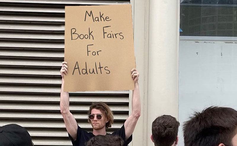 A man is holding a sign that reads ‘’Make Book Fairs For Adults.’’