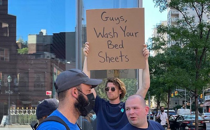 A man is holding a sign that reads ‘’Guys, Wash Your Bed Sheets.’’