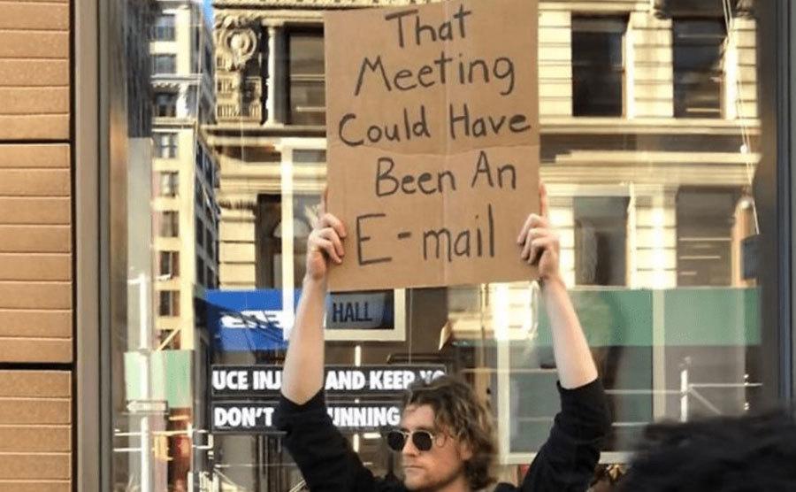 A man is holding a sign that reads ‘’That Meeting Could Have Been an Email.’’