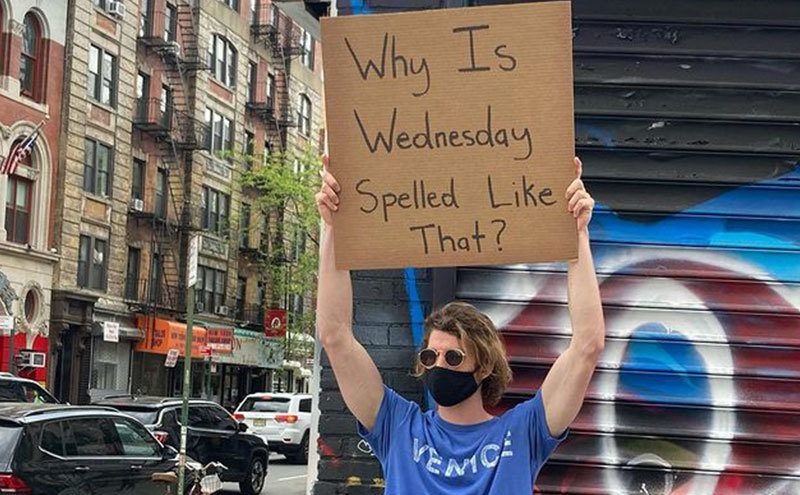 A man is holding a sign that reads ‘’Why is Wednesday Spelt Like that.’’