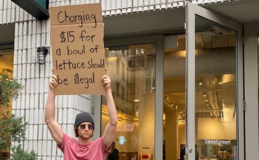 A man is holding a sign that reads ‘’Charging $15 for a Bowl of Lettuce Should be Illegal.’’