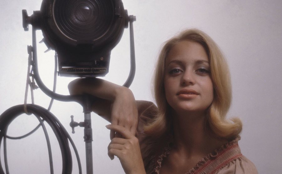Goldie Hawn, in a pink dress, leans on a studio light. 