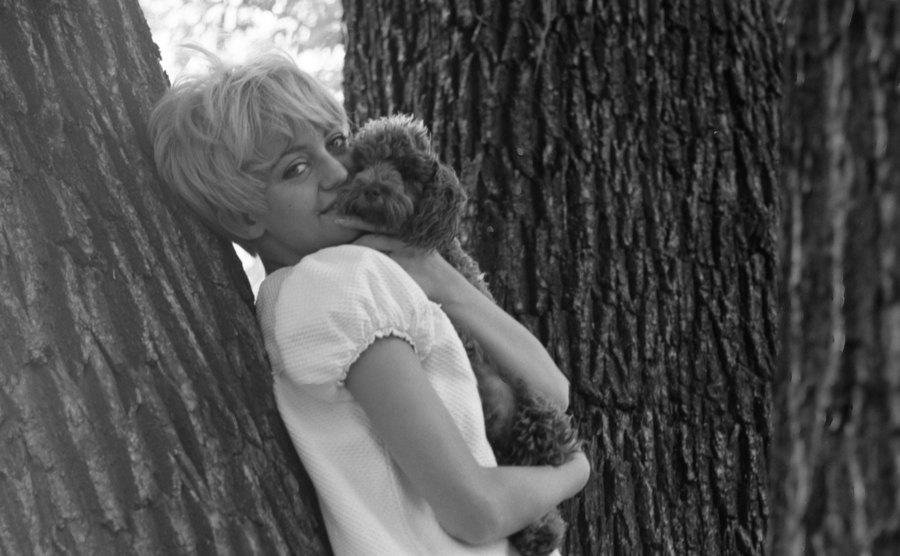 Goldie Hawn as she leans against a tree and hugs her dog. 