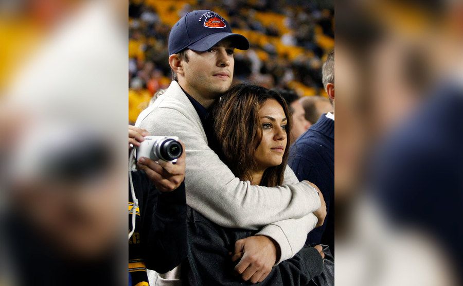 Ashton Kutcher and Mila Kunis look on from the sidelines of a basketball game. 