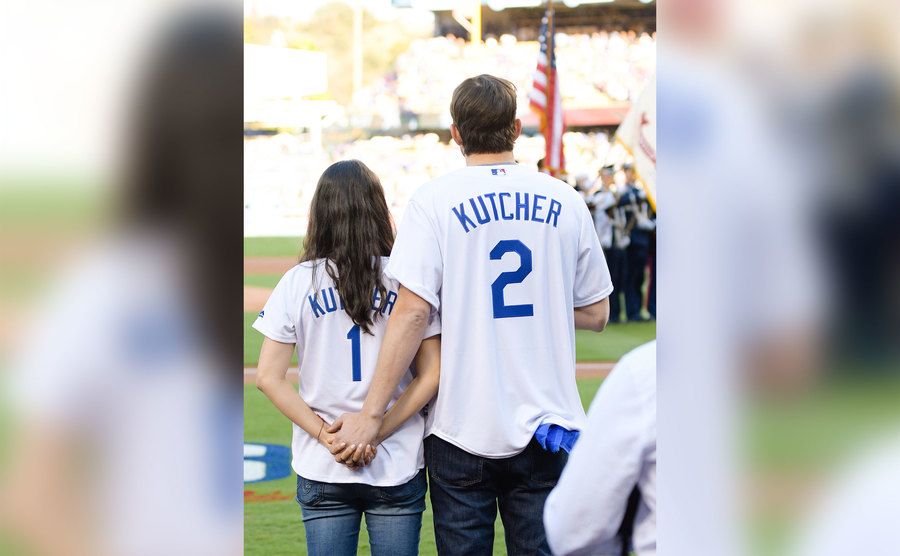 Mila Kunis and Ashton Kutcher stand as they hold hands during a baseball game. 