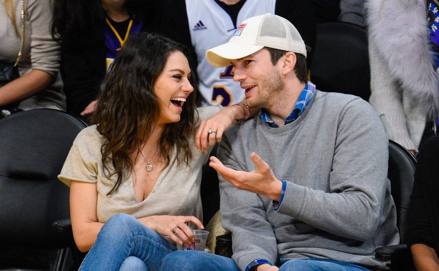 Kunis and Kutcher are laughing from the sidelines of a basketball game. 