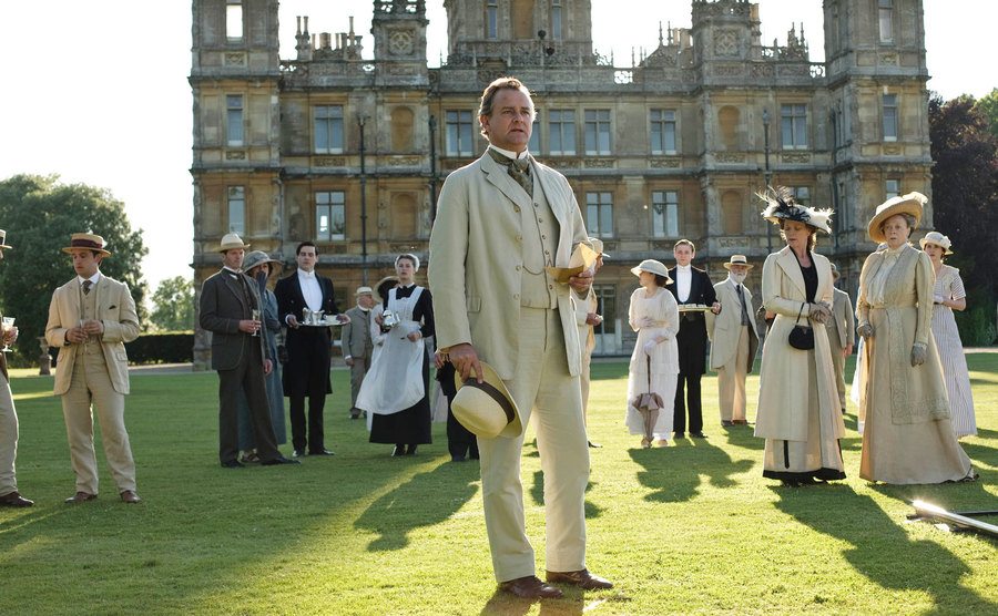 The cast of Downton Abbey stands outside on the grass, in a still from the show. 