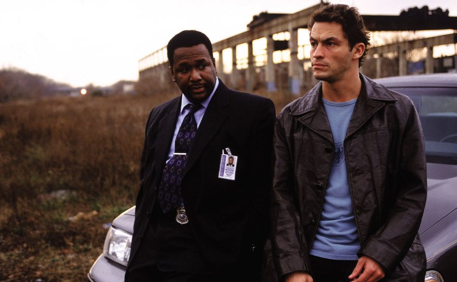 Dominic West and Wendell Pierce sit on a parked car to have a conversation. 