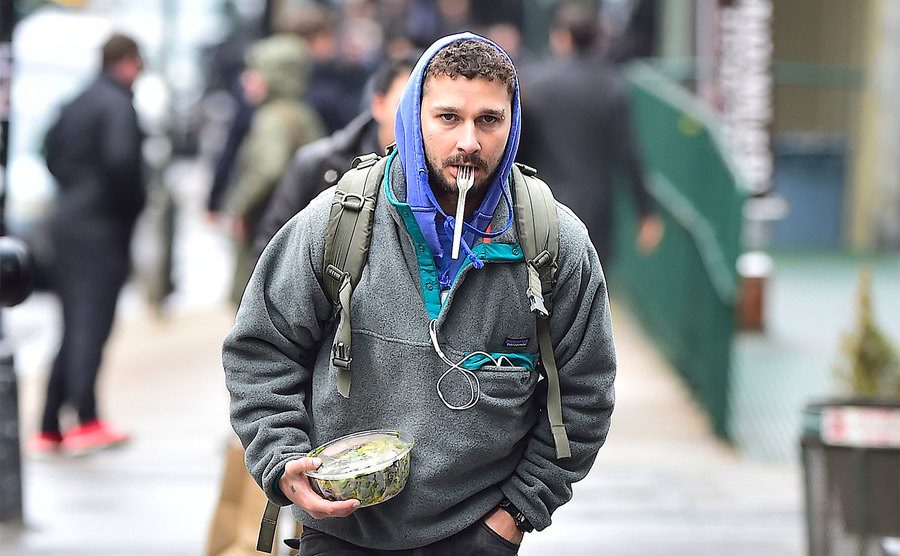 Shia LaBeouf is walking down the street with a plastic fork in his mouth. 