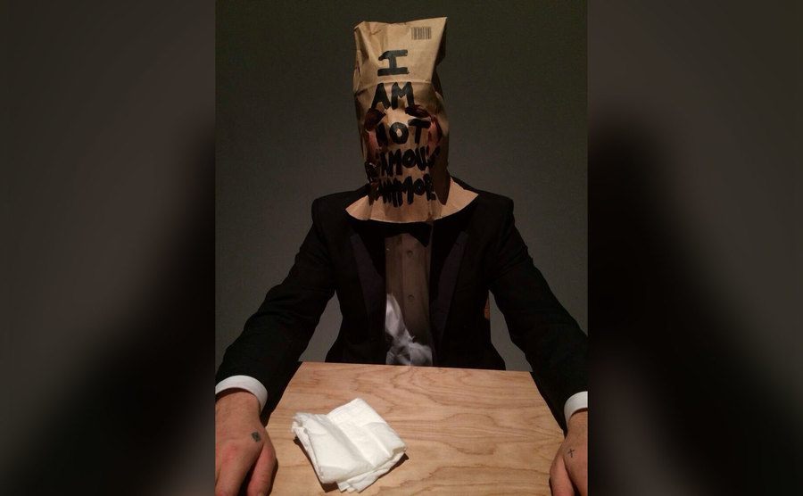 Shia sits at the table in his tux and with a paper bag over his head. 