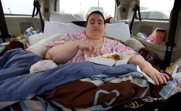 An obese woman is eating in the back of a car. 