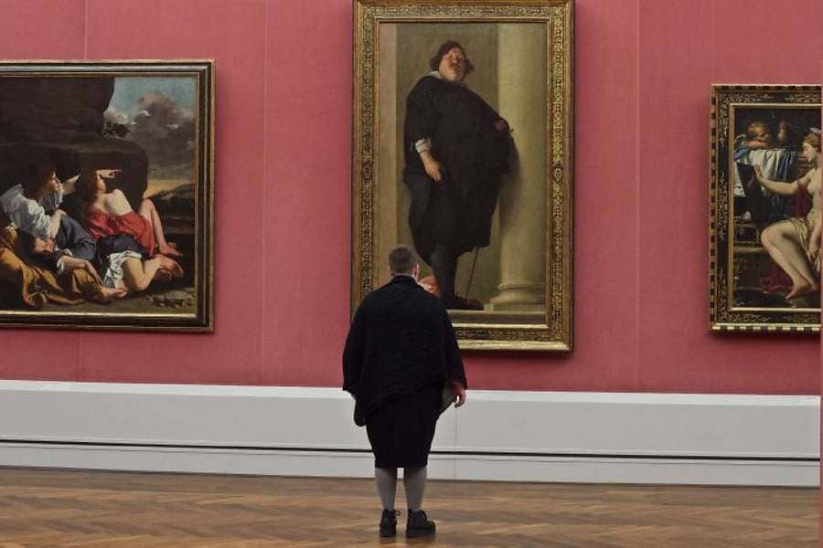 A person standing in front of a photograph of a heavy-set man in a black outfit to his knees with tights and the person wearing the same outfit.