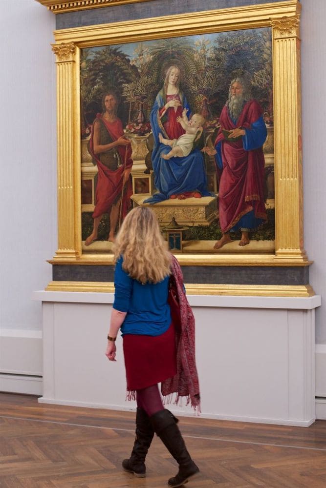 A woman walking passed a painting where both she and the people in the painting are wearing red and blue as well as a similar style.