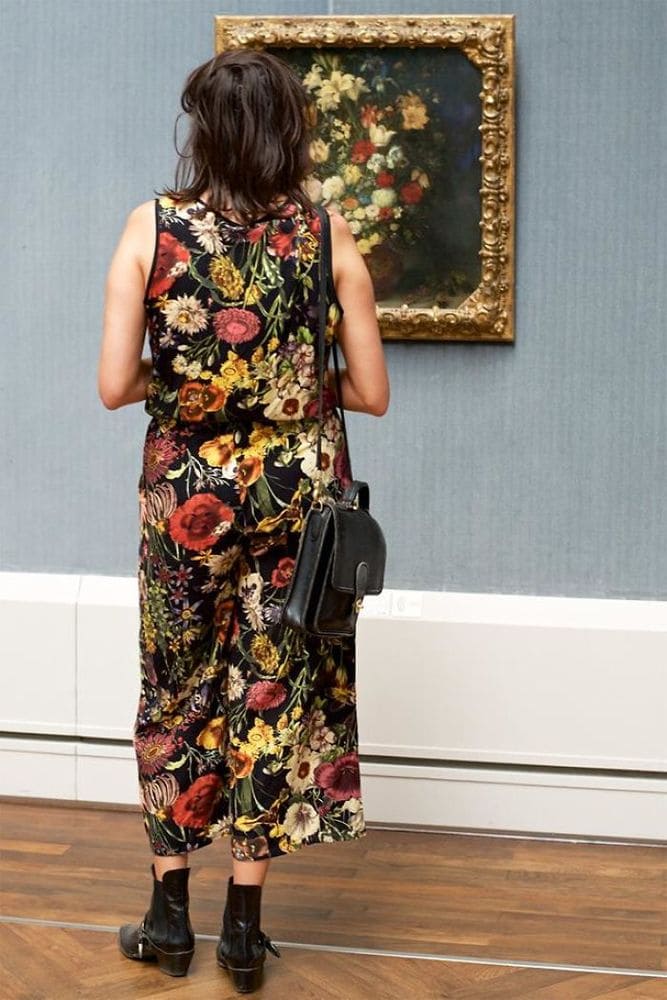 A woman with a black dress and a colorful floral pattern standing in front of a painting with a vase of flowers similar to her dress.