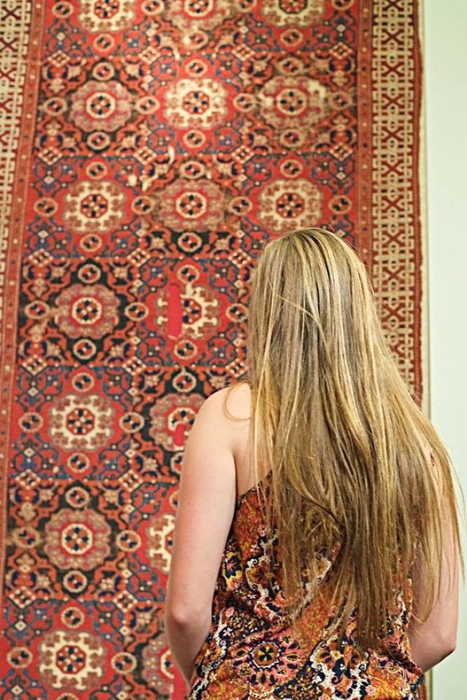 A woman wearing a patterned dress standing in front of an antique-looking carpet with a very similar pattern.