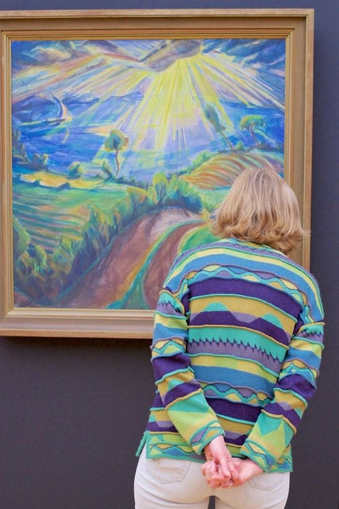 A woman in a yellow, green, and purple sweater in which the pattern matches the mountain sunset painting she is looking at.