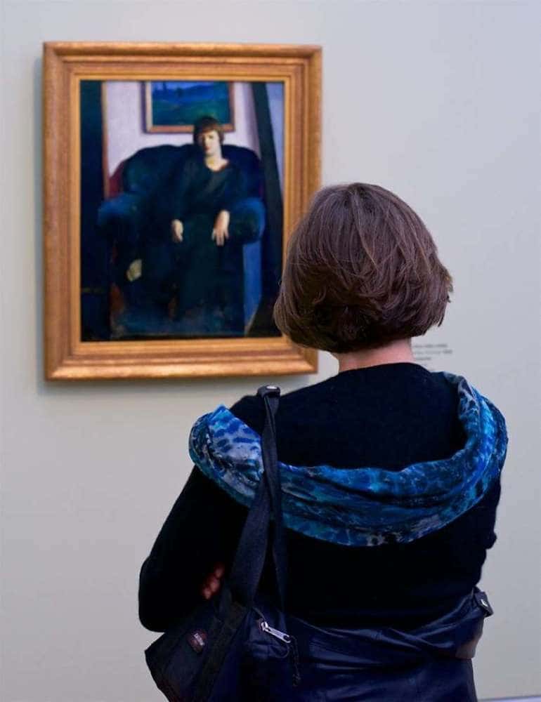 A woman with short brown hair with royal and navy blue clothing which looks like the short-haired woman sitting on a royal blue couch in the painting which she is looking at.