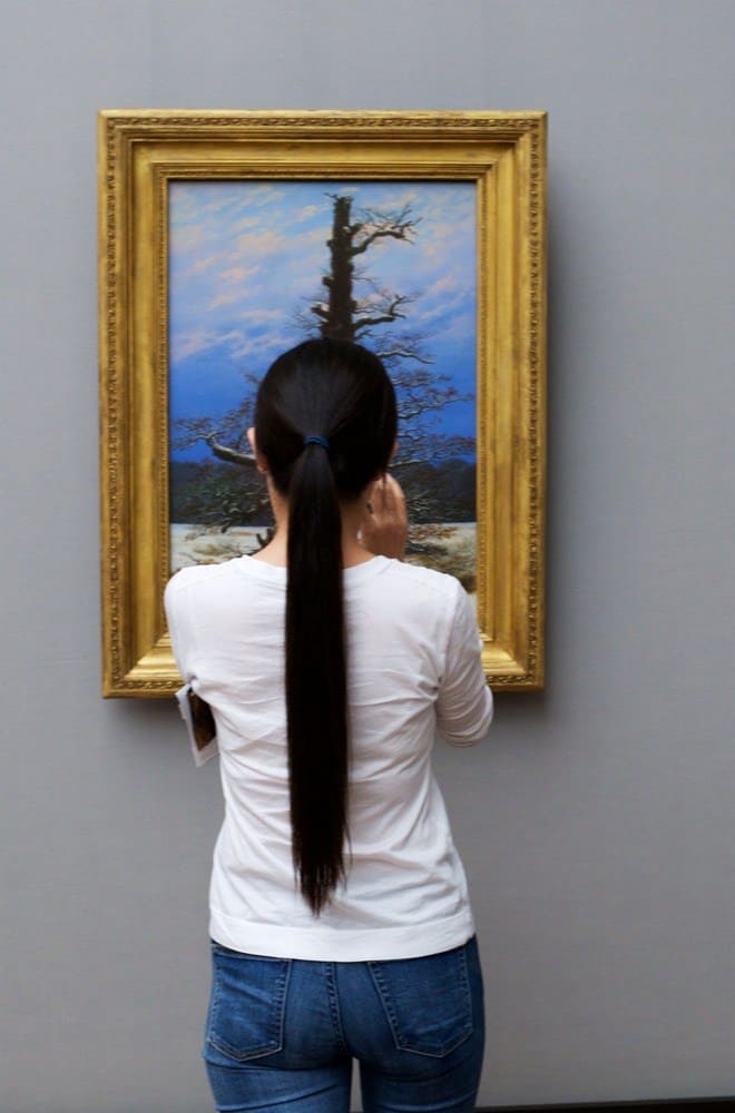A woman with a long, dark, and straight ponytail which matches the painting of a tree that she is admiring.