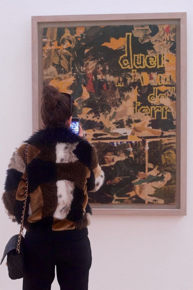A woman in a brown, black, and white faux fir coat which matches the abstract art she is photographing.