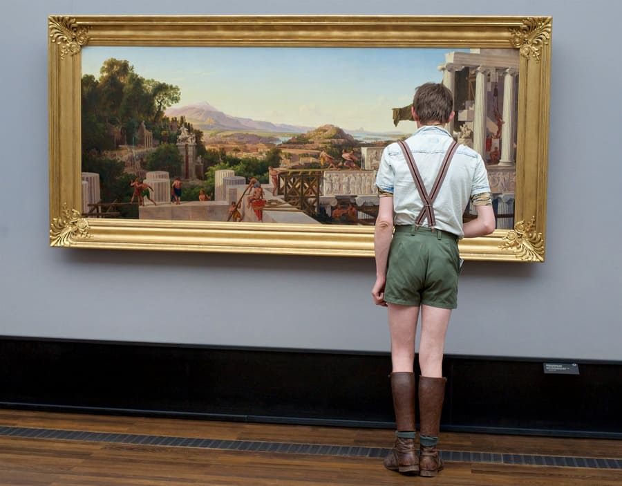 A boy in green shorts and suspenders that goes with the painting of workers building in olden times, which he is admiring.