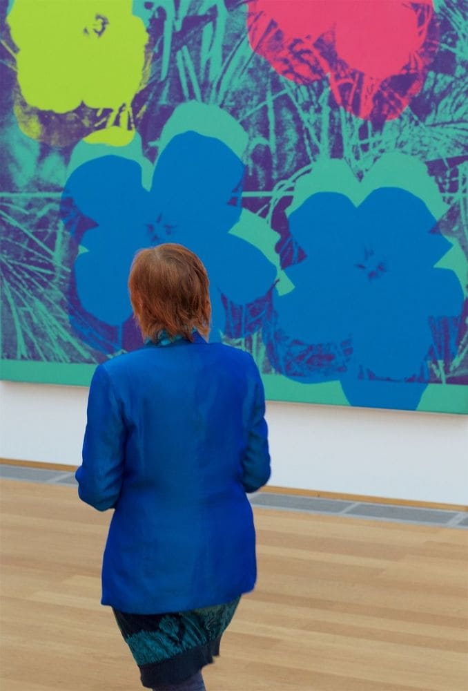A woman wearing a royal blue blazer and patterned skirt which matches the colorful floral art she is admiring.
