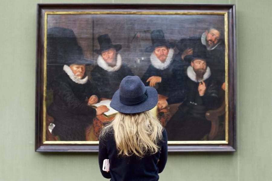 Blonde girl in a dark gray hat and black shirt admiring a similar painting of five men in black robes with tall black top hats.