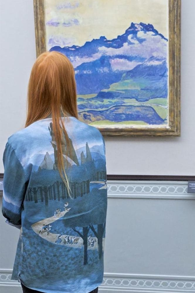 A girl wearing a shirt with a mountain and forest view on it, which matches the painting she is admiring.