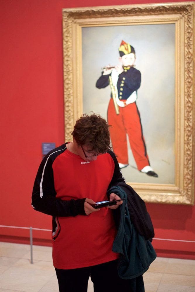 A boy on his phone wearing black and red standing in front of a painting of a boy playing the flute who is standing in the same position.