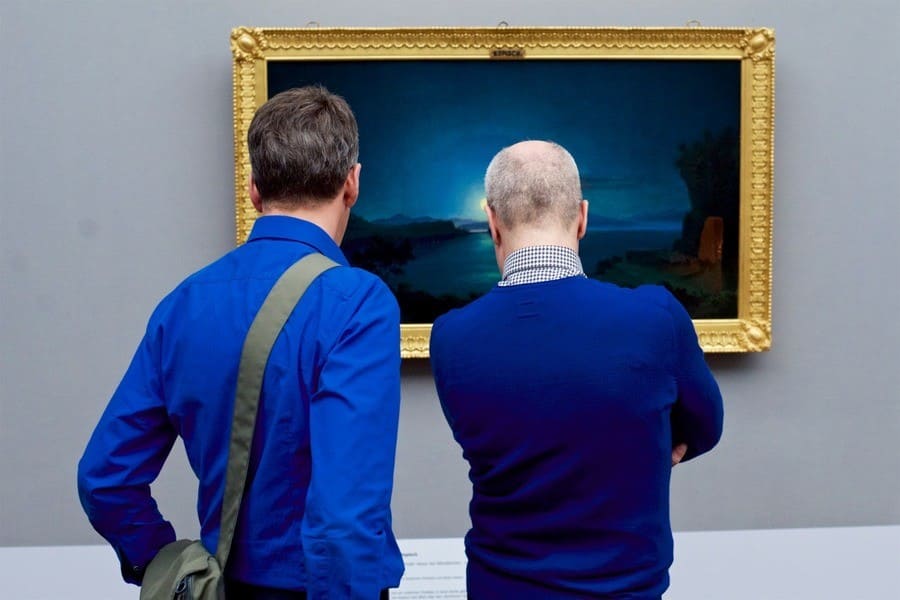 Two men in blue admiring a painting of a beautiful blue sunset.