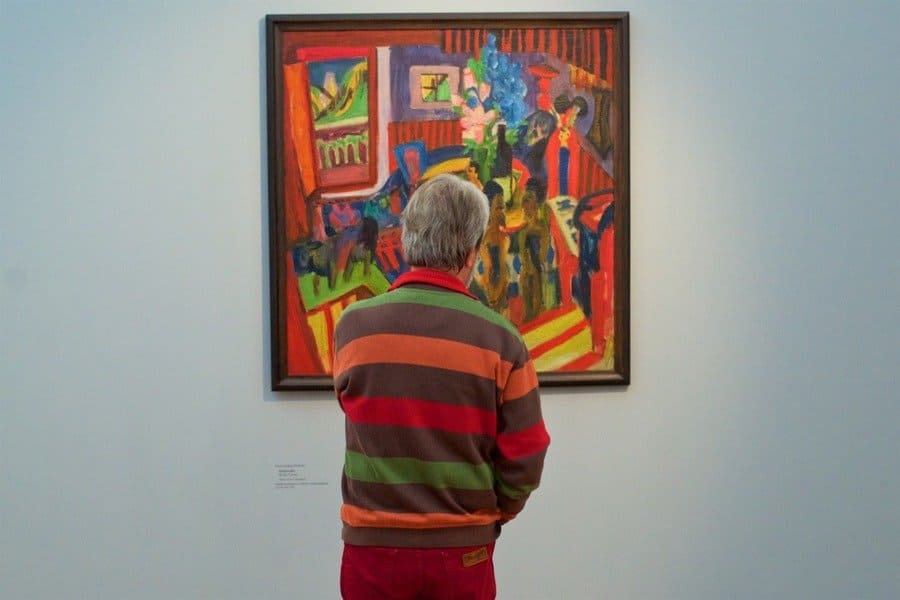 A man in a red, orange, green, and brown sweater which matches the abstract painting he is admiring.