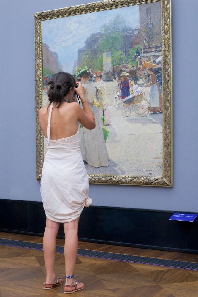 A woman wearing a white dress which matches the style of the painting she is admiring of women in the olden days wearing large dresses.