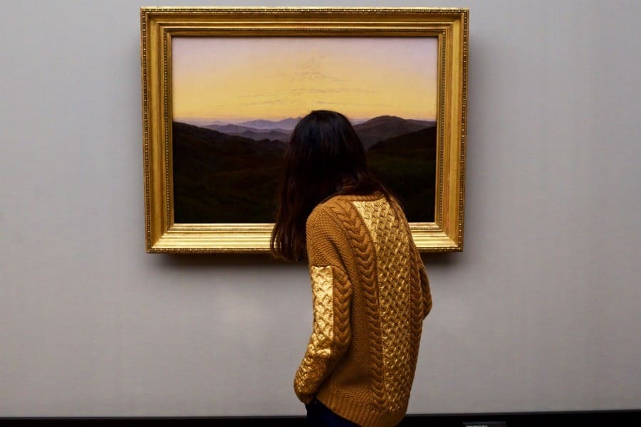 Woman wearing a gold sweater that matches the frame and part of the sunset of the painting she is admiring.