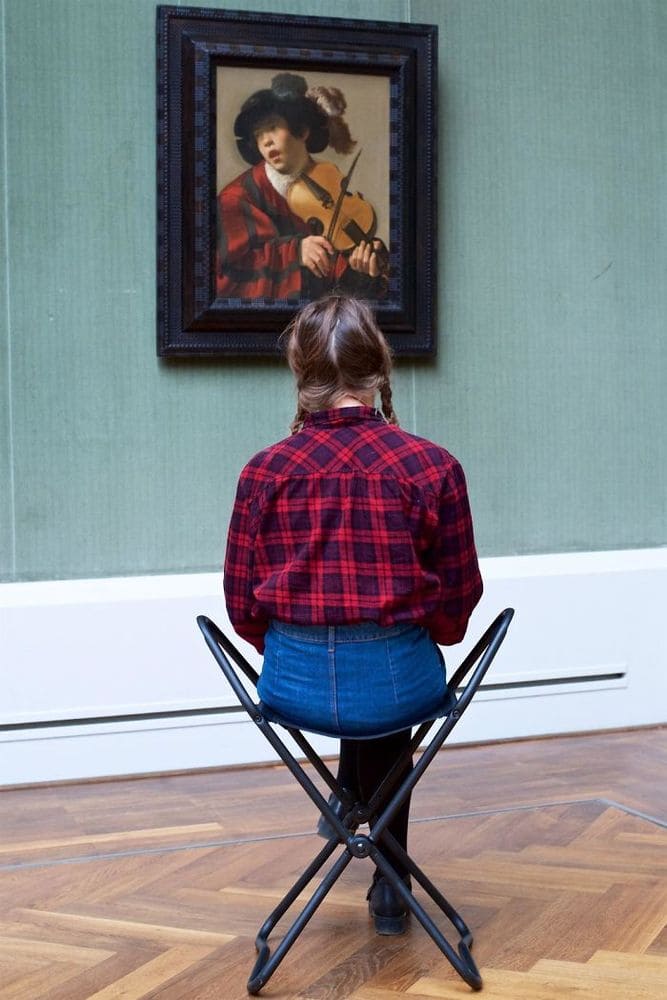 Woman in a red plaid shirt sitting in front of a painting of a person playing the violin in a similar shirt.
