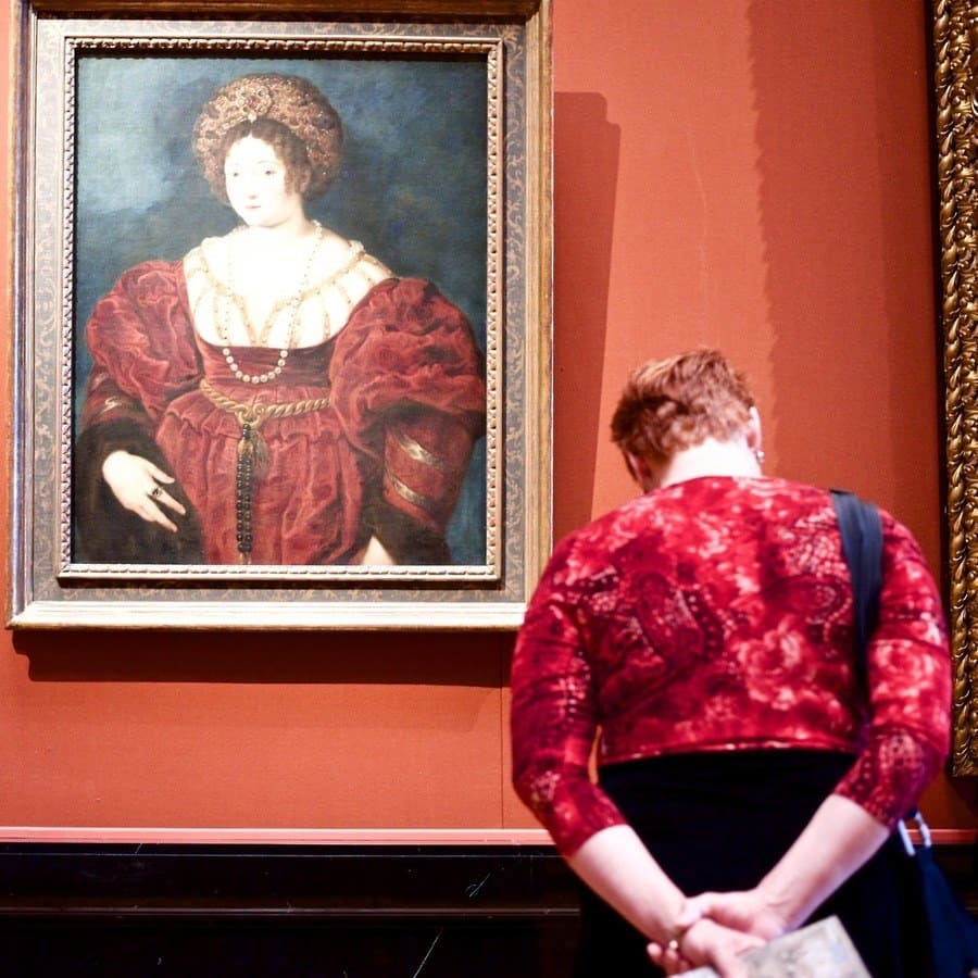 Painting of a Victorian woman wearing a dark red dress that matches the clothing of the lady admiring it.