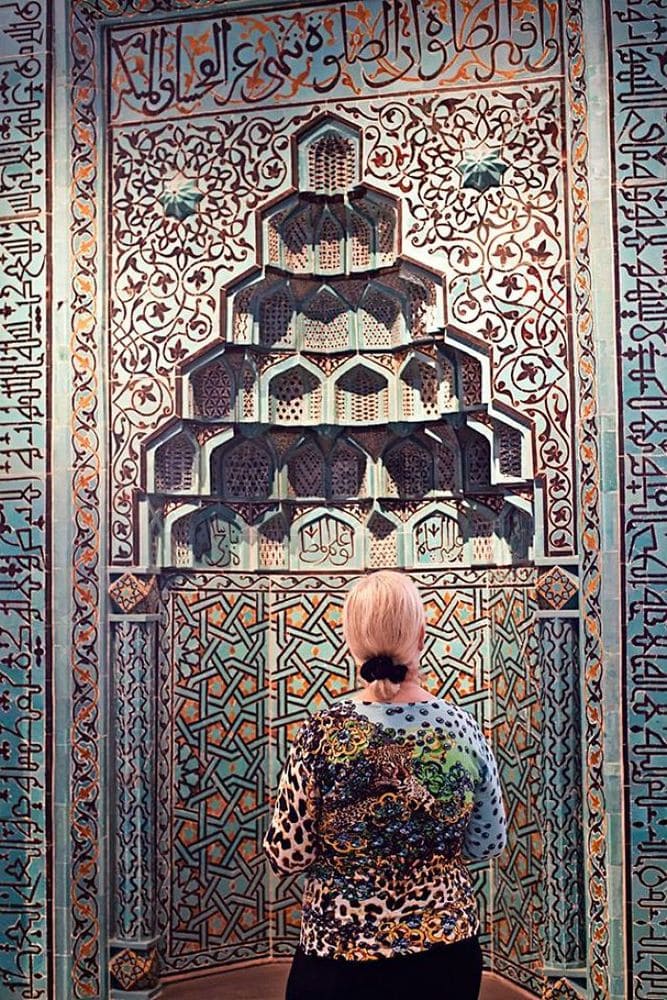 A wall of tiled light blue, orange, and brown artwork being admired by a woman wearing a shirt with similar colors and patterns.