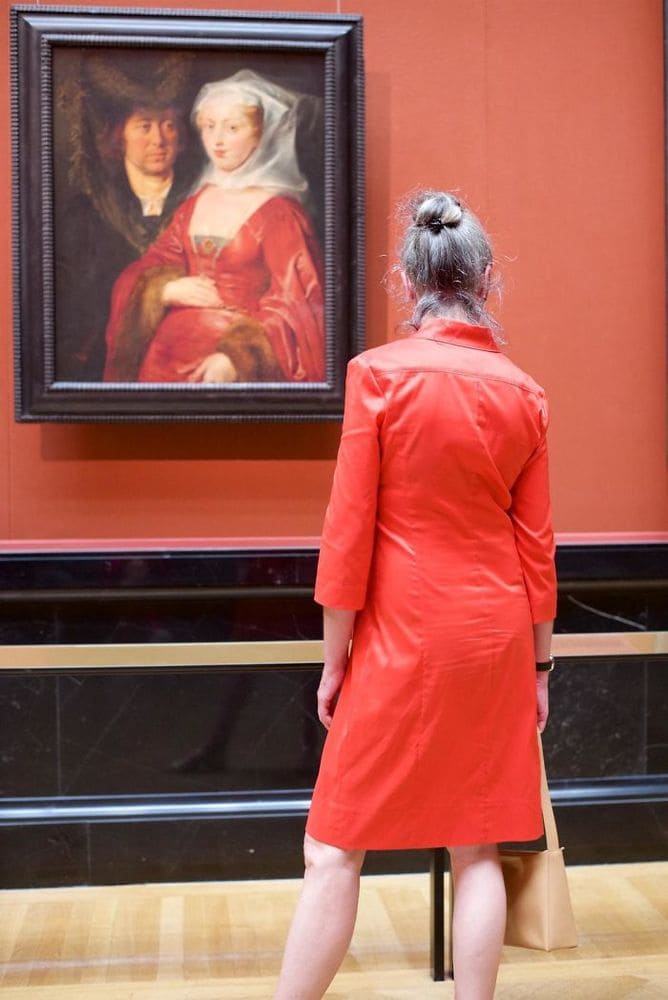 Woman in a red dress standing in front of a painting of a couple, where the woman is also wearing a red dress.