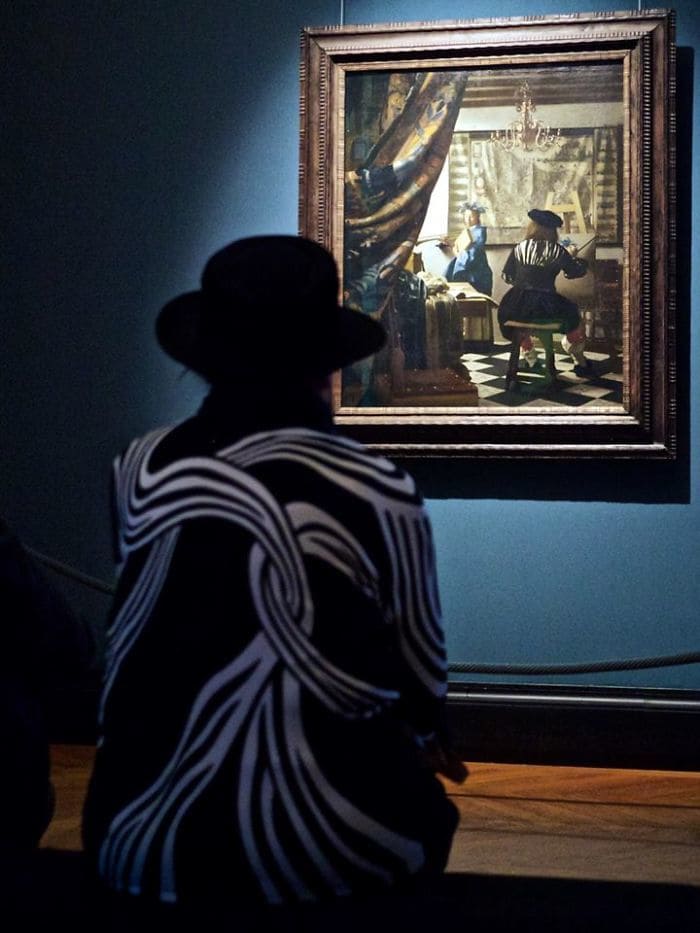 A woman in a black hat and a black sweater with some white design on it admiring a painting of someone who looks similar painting on a canvas.