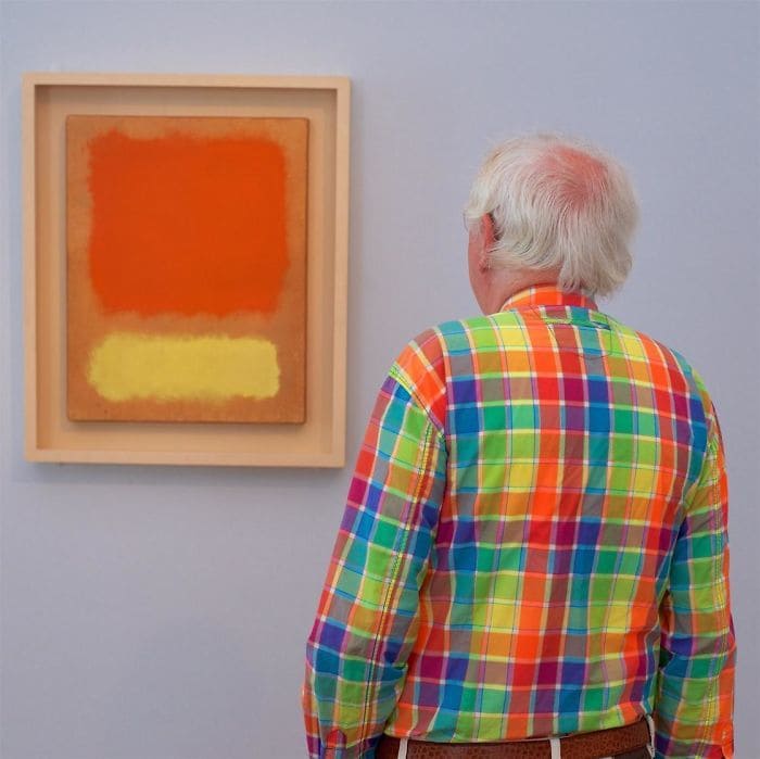 An abstract painting in yellow and orange, which matches the multi-colored square-patterned shirt of the man admiring it.