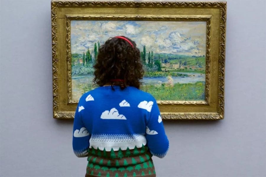 A woman wearing a sweater with clouds and a field of trees on it while admiring a painting of fields and trees.