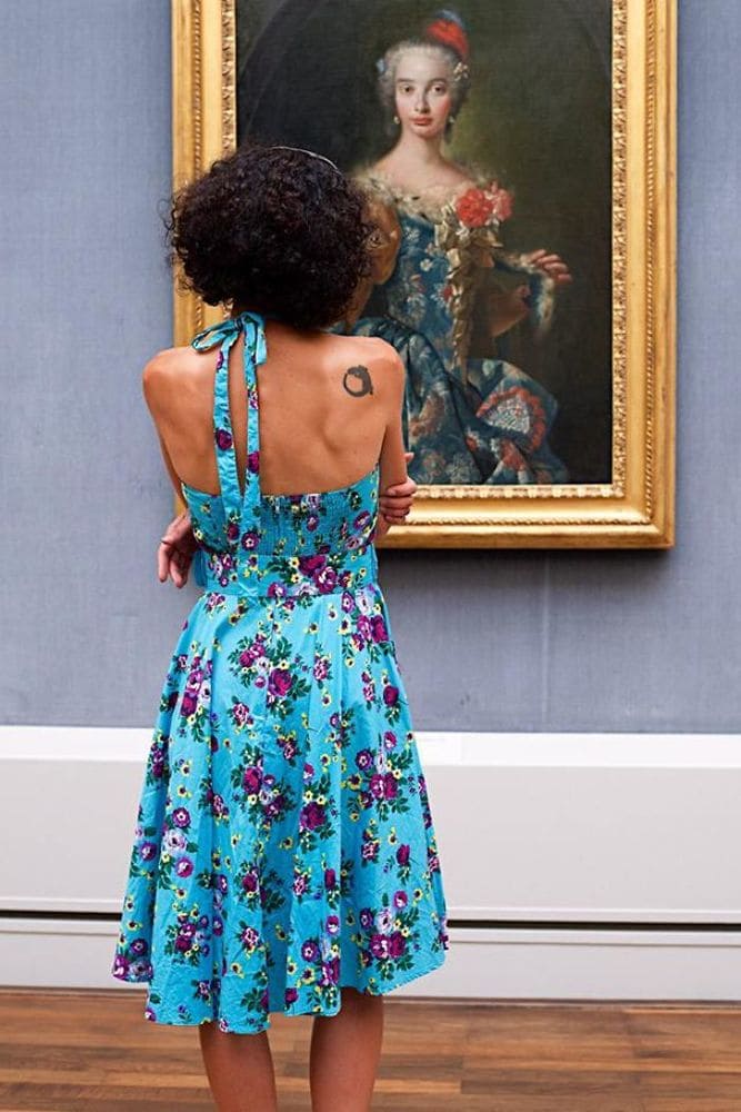 A woman wearing a blue floral patterned dress admiring a painting of a woman who is also wearing a blue floral dress.